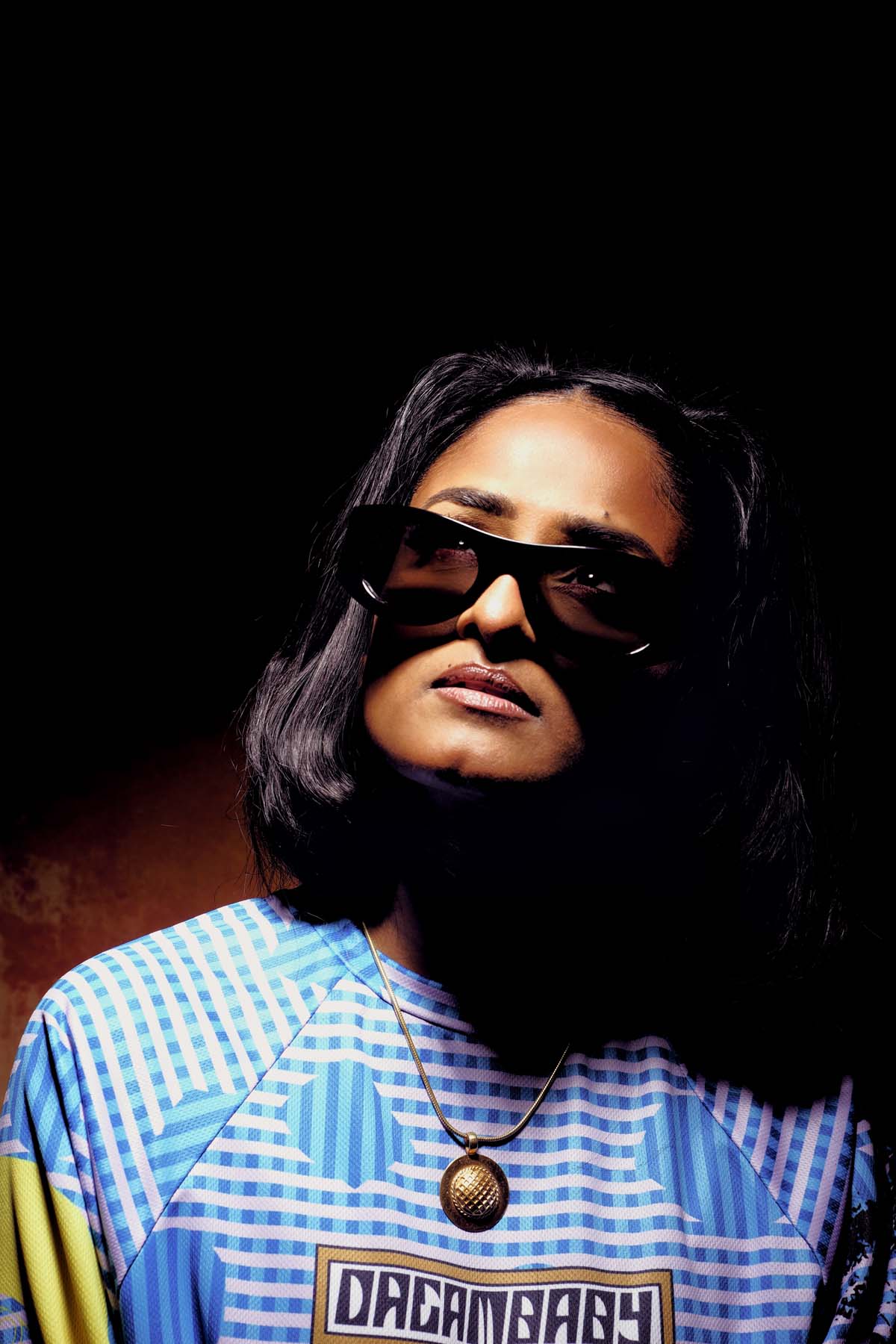 A woman with black shoulder-length hair can be seen. Priya Ragu is wearing sunglasses, her eyes are visible through the lenses. She as is illuminated from above, the background is dark. She is looking upwards, wearing a colourful shirt with the word "Dreambaby" and a gold necklace with a round pendant.