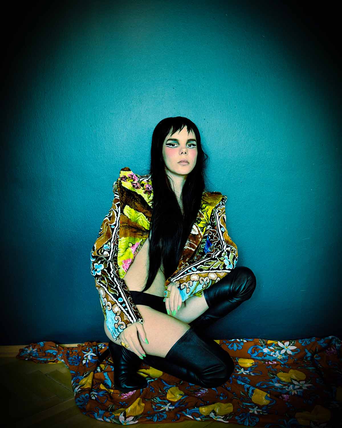Young woman dressed in black panties, black overknee boots and a colourful, wildly patterned blazer squats in front of a turquoise wall on a flower-printed fabric. Gaye Su Akyol's long black hair covers her upper body, which is seemingly naked under the jacket. Her eyes are strikingly made up, she wears long poison green fingernails and looks serious into the camera.
