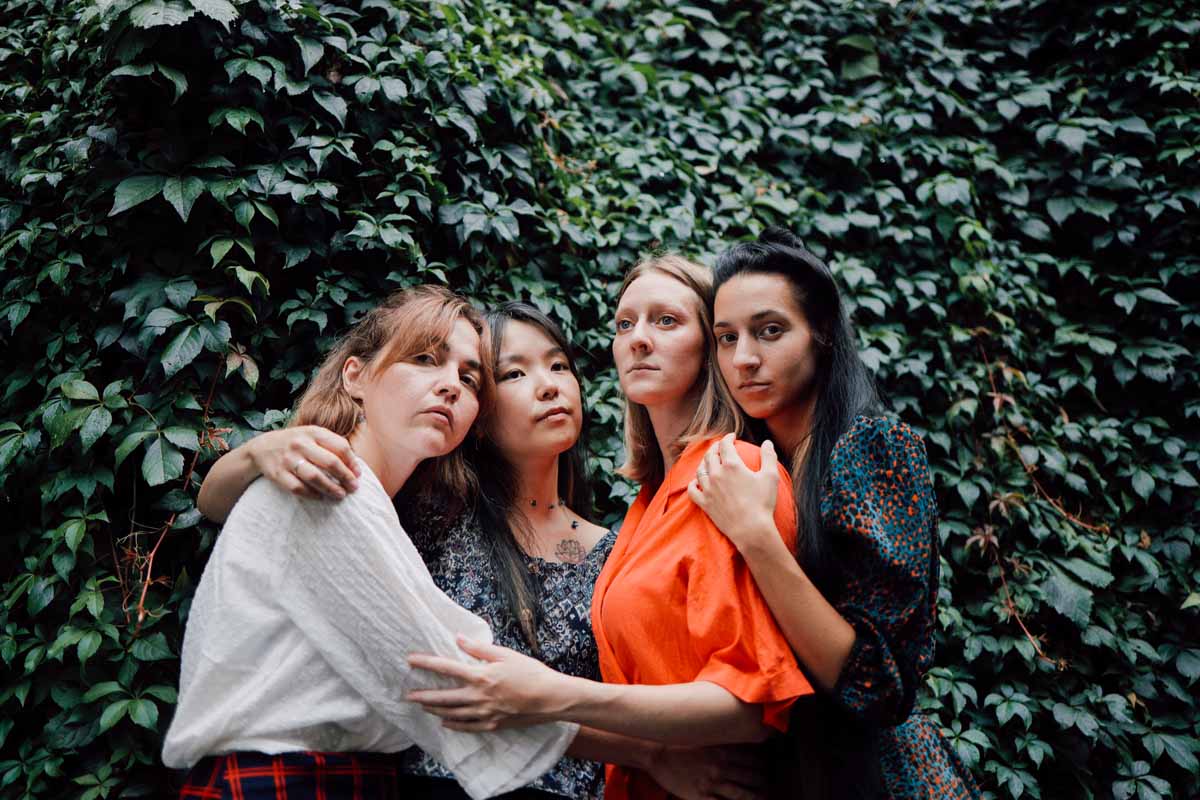 Four young women stand in front of a green hedge that fills the background of the picture, gently touching each other - sometimes with their arms around each other or on each other's shoulders and thus connected. The members of LUCIDVOX look in different directions, ranging from expressionless to melancholic.