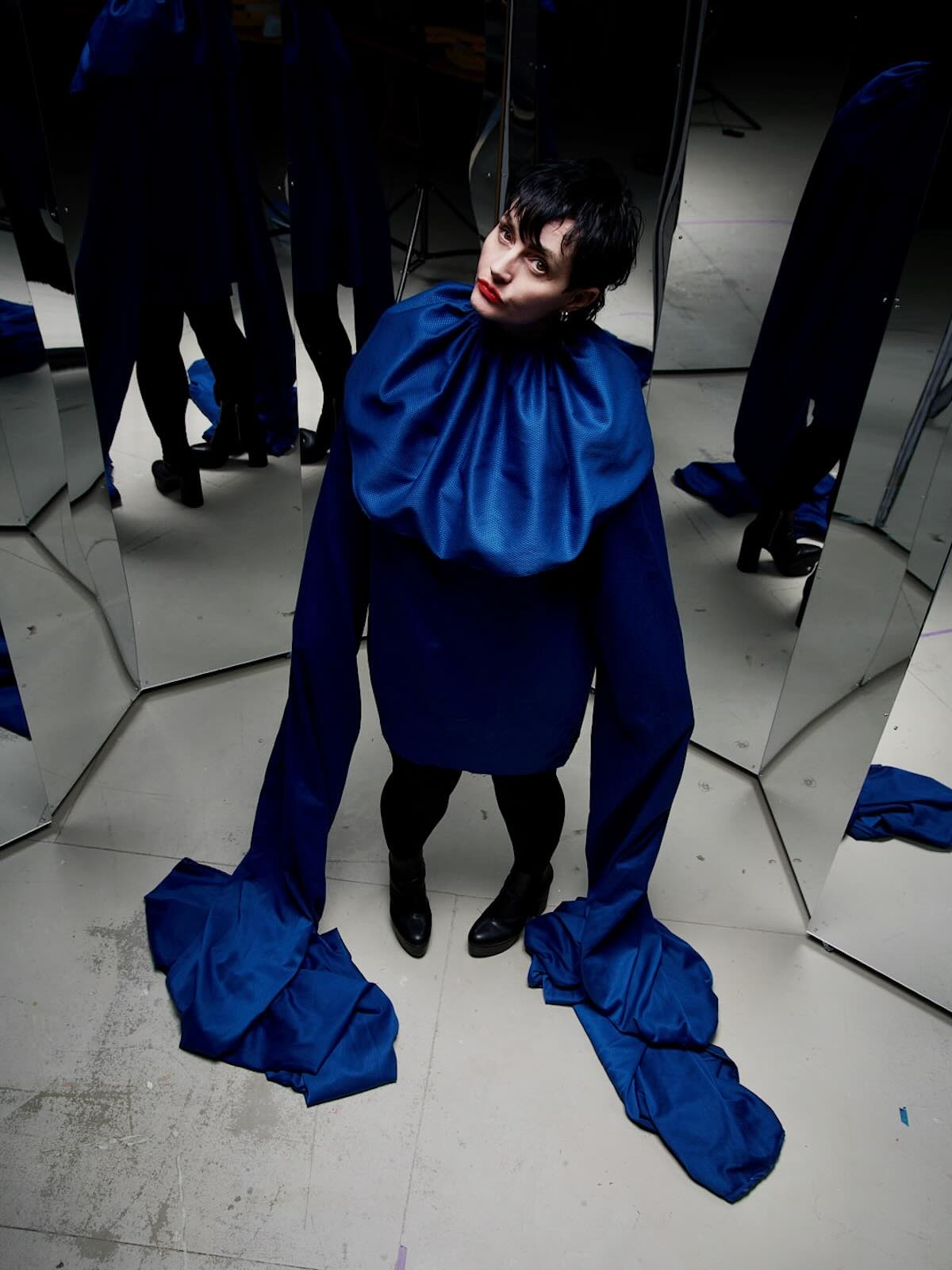 The image shows Melanie Jame Wolf standing in front of several mirrors, creating multiple reflections. She is wearing a striking blue outfit with a high collar and long, flowing sleeves that drape to the floor. Her short dark hair and red lipstick contrast with the blue fabric, and she is looking slightly upward with a contemplative expression. The setting appears to be an artistic or fashion photoshoot, with a neutral, industrial background.