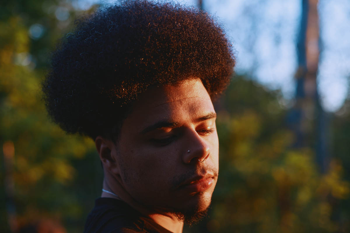 Close-up of the head of a Black person looking down thoughtfully. The evening sun shines on the side of Otis Mensah's face. Otis Mensah wears an Afro, a nose stud and a septum ring.