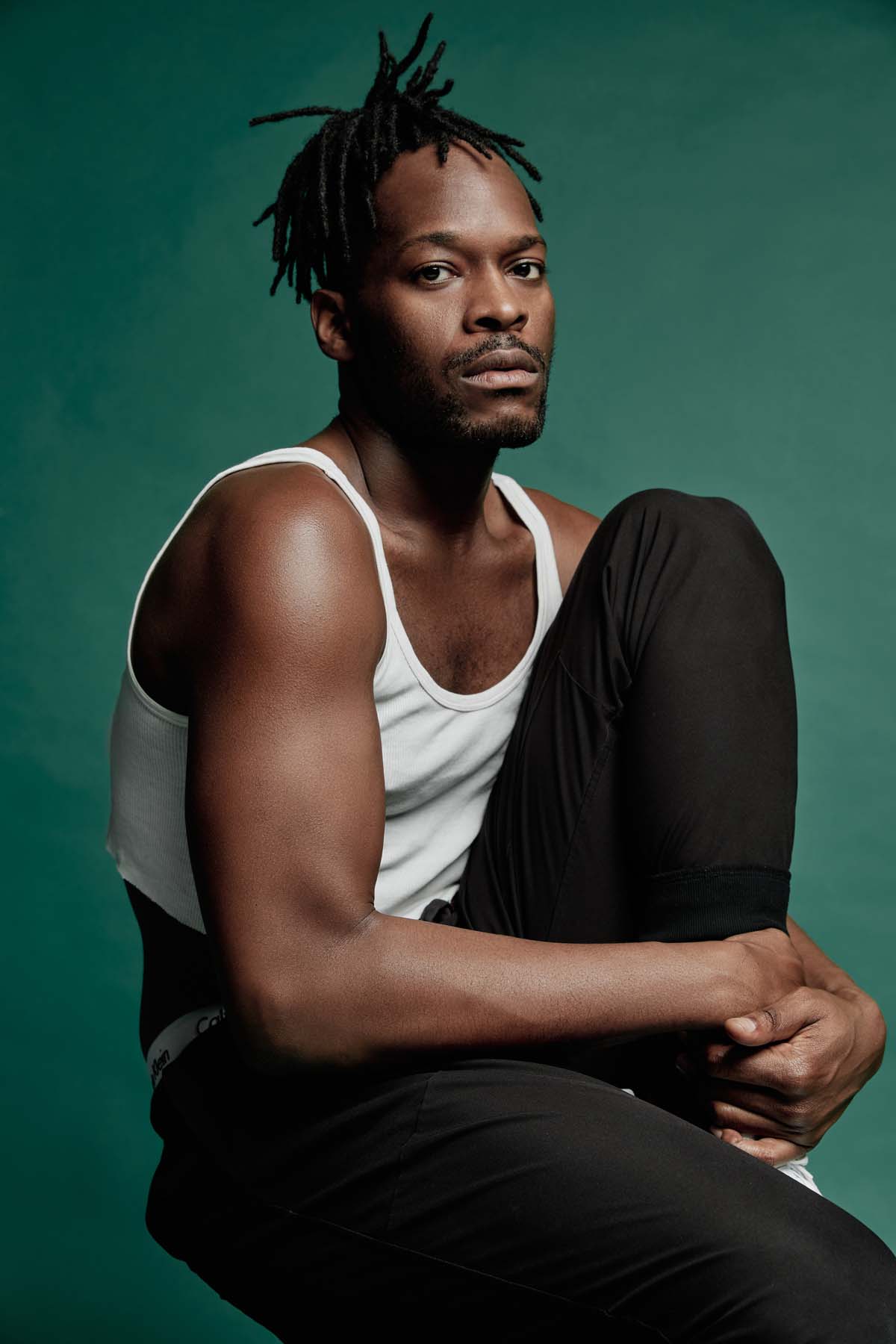 A Black male sits on a stool, has one leg bent on it and clasps his shin with both hands. Zebra Katz wears black, short dreadlocks, a Henriquatre beard and looks sternly into the camera. He is wearing a belly-free white tank top and black trousers.