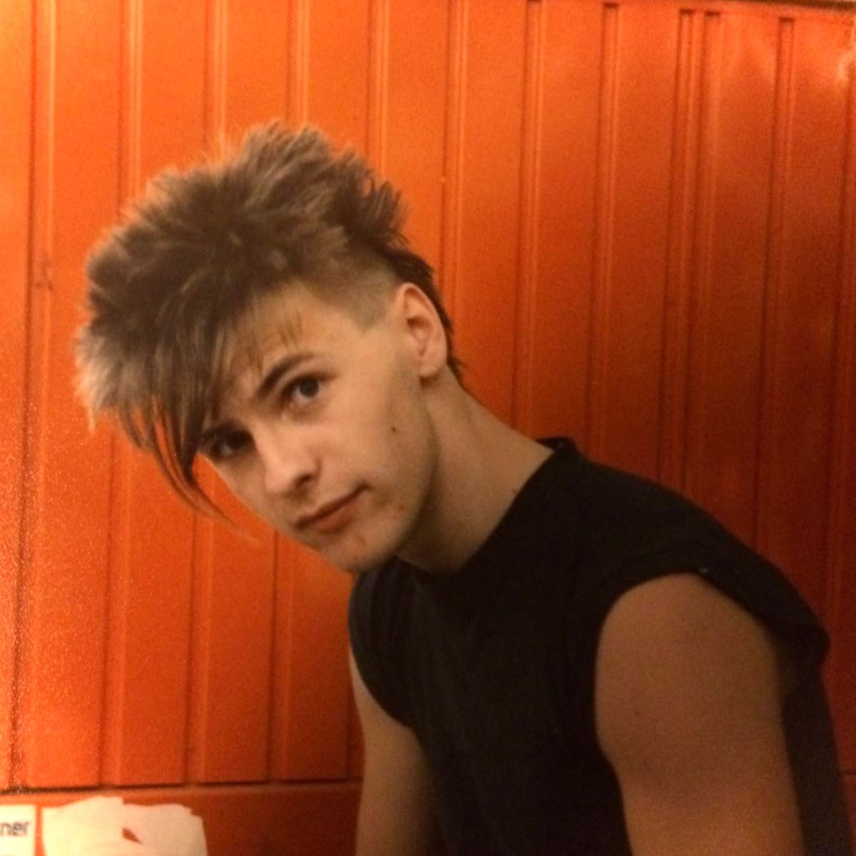 The image shows a young Ulrich Gutmair in front of an orange wall. He has a distinctive hairstyle with spiky, wild hair and shaved sides. He is wearing a sleeveless black shirt and is looking slightly down at the camera with an attentive and serious expression.