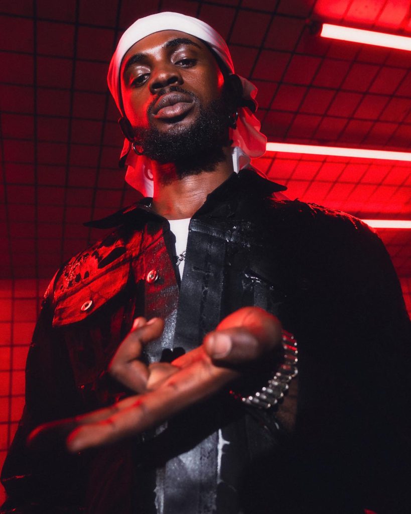 The image shows Black Sherif standing in a dimly lit environment with red neon lights. He is wearing a black jacket and a white bandana on his head. He looks confidently into the camera, with one hand extended towards the viewer, partially in focus. The background features a grid pattern on the ceiling, adding a modern and edgy atmosphere to the photo. The red lighting casts dramatic shadows, highlighting his facial features and creating a striking visual effect.