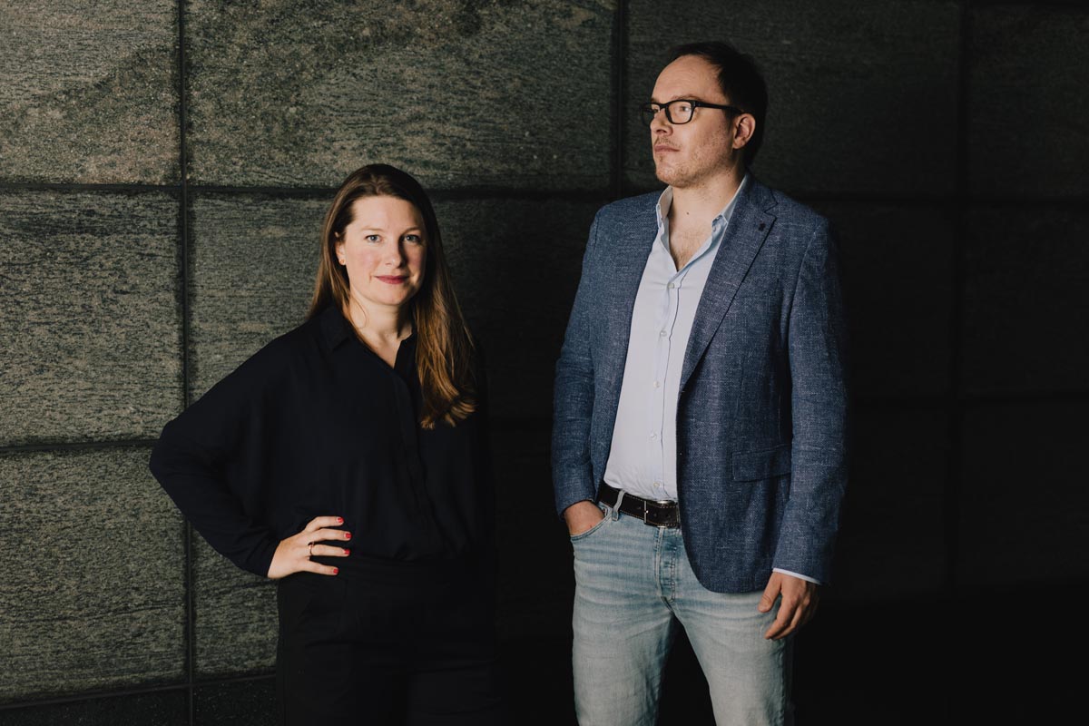 Das Bild zeigt Lena Kampf und Daniel Drepper. Lena Kampf steht links und trägt ein schwarzes Oberteil, während Daniel Drepper rechts steht und ein blaues Sakko mit einem hellblauen Hemd und Jeans trägt. Sie stehen vor einem dunklen, strukturierten Hintergrund. Lena Kampf hat ihre rechte Hand auf der Hüfte und lächelt leicht in die Kamera. Daniel Drepper hat seine Hände in den Hosentaschen und blickt nach rechts. Die gesamte Szene wirkt professionell und ernst.