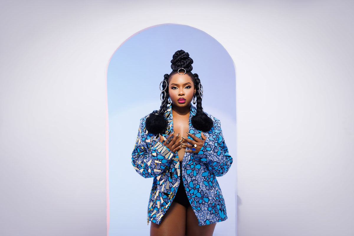 The picture shows Yemi Alade standing in front of a white and light blue arched backdrop. Her hair is styled in an intricate updo with large braids and circular hair accessories. She is wearing a blue patterned blazer with floral designs, slightly open to reveal a black outfit underneath. Her makeup is bold, featuring bright lipstick, and she has large hoop earrings. Her hands, adorned with multiple rings, are raised to her chest, giving a confident and stylish pose. The minimalist background draws focus to her striking appearance and attire.