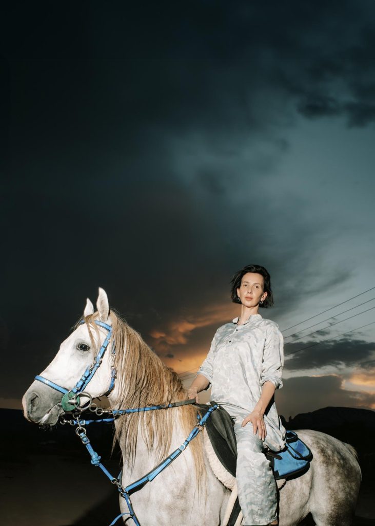 Das Bild zeigt Zuzanna Czebatul, die auf einem weißen Pferd sitzt und die Zügel hält. Sie trägt ein helles, gemustertes Outfit und blickt mit einem ruhigen Ausdruck direkt in die Kamera. Im Hintergrund ist ein dramatischer, bewölkter Himmel mit einem Hauch von Sonnenuntergang zu sehen, was der Szene Tiefe und Atmosphäre verleiht.