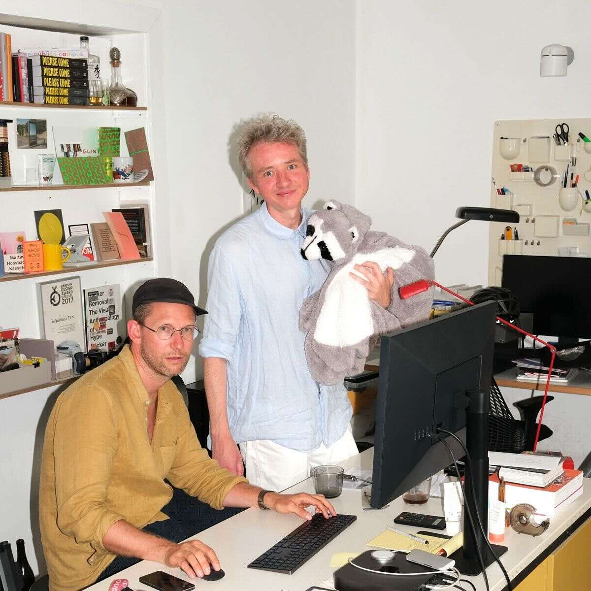 In the image, Norman Palm and Alexander Winkelmann from Fertig Design are seen. Norman Palm is sitting at a desk in front of a computer, wearing glasses and a black cap. Alexander Winkelmann is standing next to him, holding a large plush raccoon. In the background, there are shelves with books and various items. They are in a well-lit office.