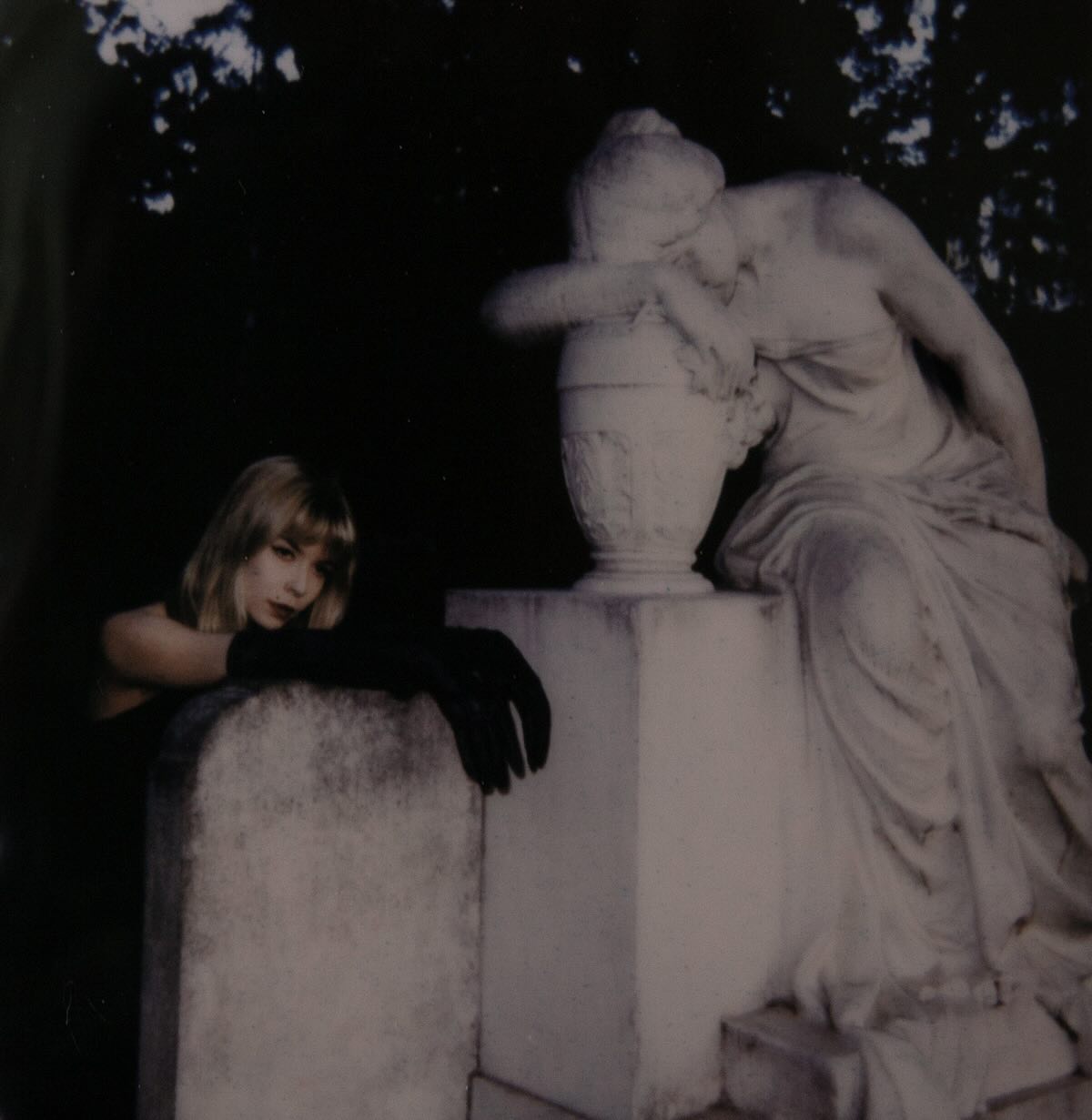 Anni Porrasmäki, dressed in black with long gloves, leans on a gravestone next to a statue of a weeping woman in a dark, moody setting. The statue is leaning on an urn, and the overall atmosphere is somber and artistic.