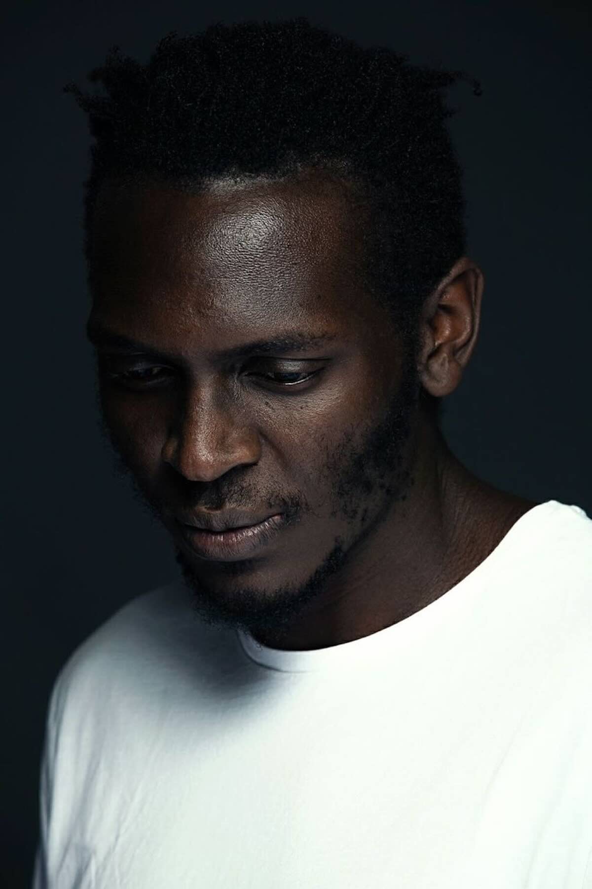 Portrait of Emo Rugene. He is wearing a white T-shirt and looking down. The image has a dark background and conveys a contemplative mood.