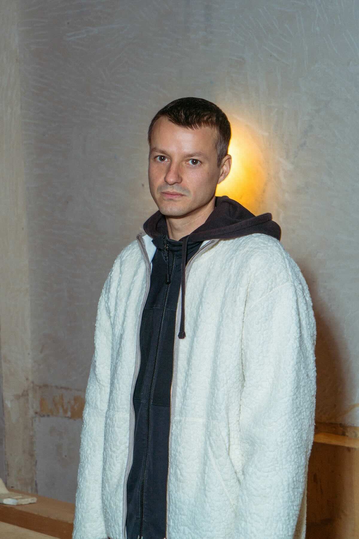 In the image, Levente is seen standing indoors against a neutral background with soft lighting. He is wearing a white, textured jacket over a dark hoodie. Levente has short hair and is looking directly at the camera with a neutral expression.